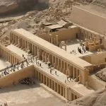 An aerial view of the terraced structure of the Temple Of Hatshebsut, nestled into the dramatic cliffs of Deir el-Bahari in Luxor, Egypt, with columns and courtyards visible.