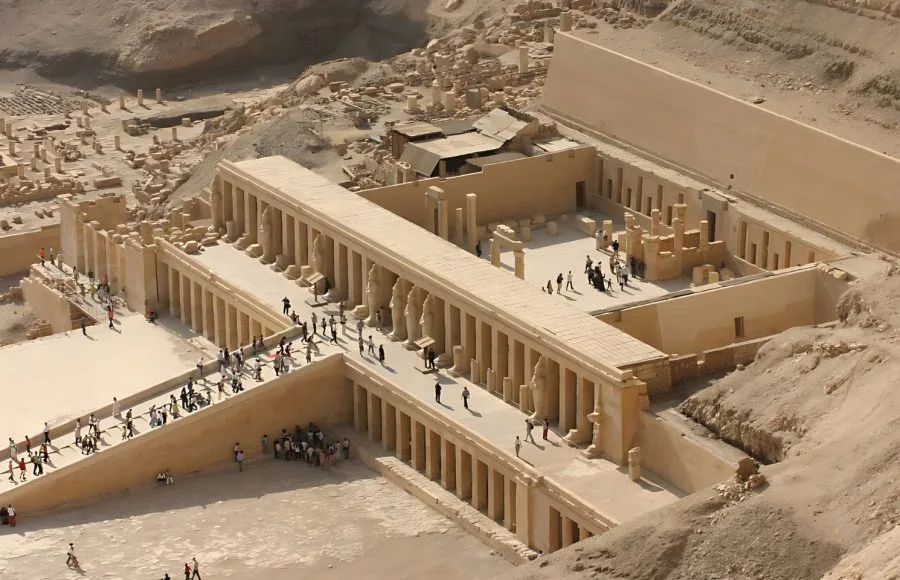 An aerial view of the terraced structure of the Temple Of Hatshebsut, nestled into the dramatic cliffs of Deir el-Bahari in Luxor, Egypt, with columns and courtyards visible.