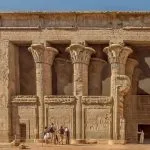 Tourists standing before the ornate entrance of the Temple of Esna with a Hall of 24 Pillars Dedicated to the God Khnum, highlighting the massive stone columns and detailed hieroglyphic reliefs.
