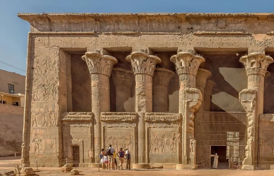 Tourists standing before the ornate entrance of the Temple of Esna with a Hall of 24 Pillars Dedicated to the God Khnum, highlighting the massive stone columns and detailed hieroglyphic reliefs.