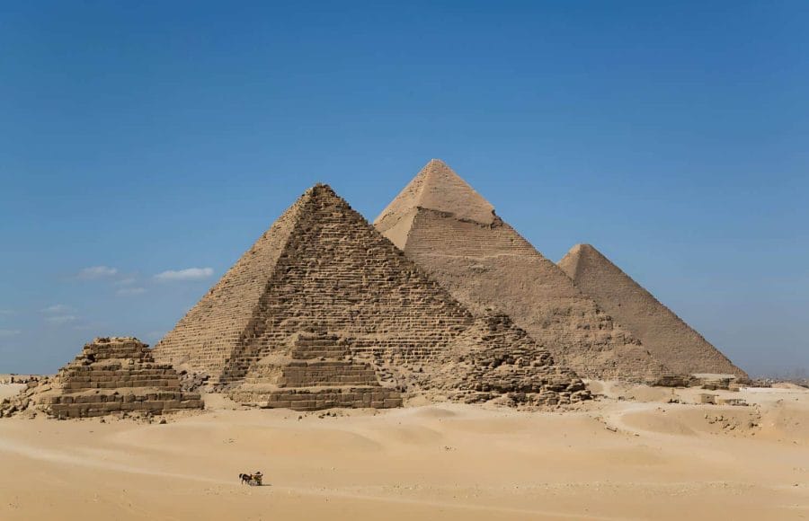 A classic wide-angle view of the three main Giza Pyramids and smaller queen's pyramids, set against the clear blue sky, with a small horsecart visible in the foreground desert sand.