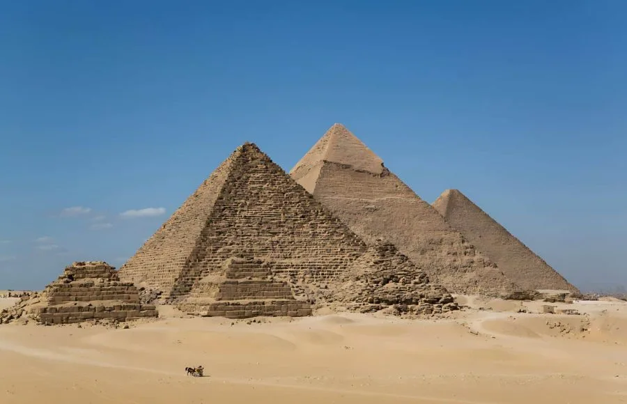 A classic wide-angle view of the three main Giza Pyramids and smaller queen's pyramids, set against the clear blue sky, with a small horsecart visible in the foreground desert sand.