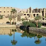 View of the Sacred Lake at Karnak Temple with palm trees reflecting in the water and the temple ruins in the background.