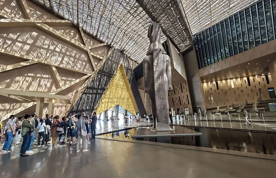 Tourists viewing the colossal statue in the stunning, sunlit atrium of the new Grand Egyptian Museum in Giza.