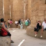Tourists relax on stone benches in the Great Hypostyle Hall of Karnak Temple, surrounded by towering carved columns and hieroglyphs.