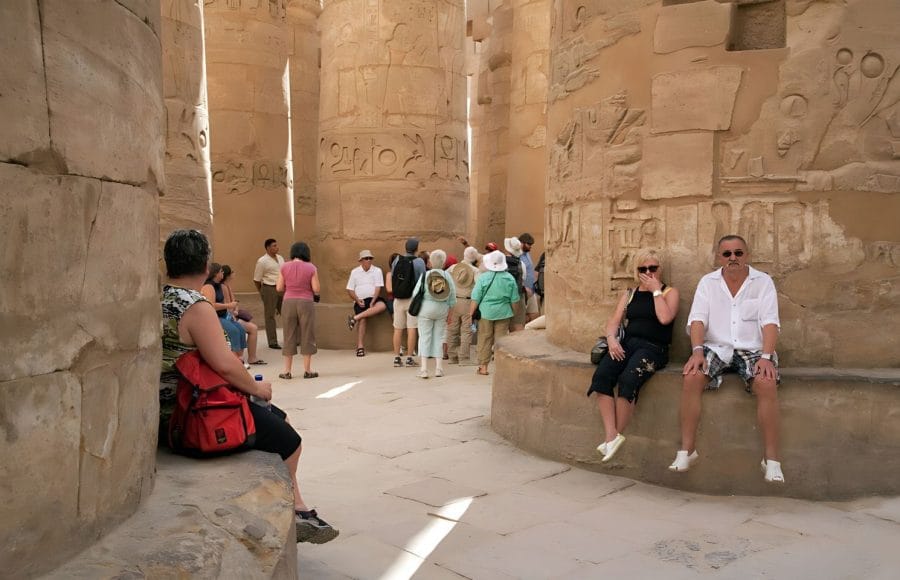 Tourists relax on stone benches in the Great Hypostyle Hall of Karnak Temple, surrounded by towering carved columns and hieroglyphs.