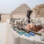 A foreground display of Egyptian souvenirs (scarabs, pyramids, Nefertiti busts) with Tourists with the Saqqara Step Pyramid of Djoser visible in the background against the desert landscape.