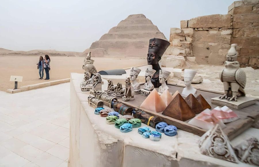 A foreground display of Egyptian souvenirs (scarabs, pyramids, Nefertiti busts) with Tourists with the Saqqara Step Pyramid of Djoser visible in the background against the desert landscape.