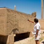 A tourist taking photos of Hapshetsut granite, specifically an engraved, massive block featuring detailed hieroglyphic reliefs and an obelisk in the background at Karnak Temple.