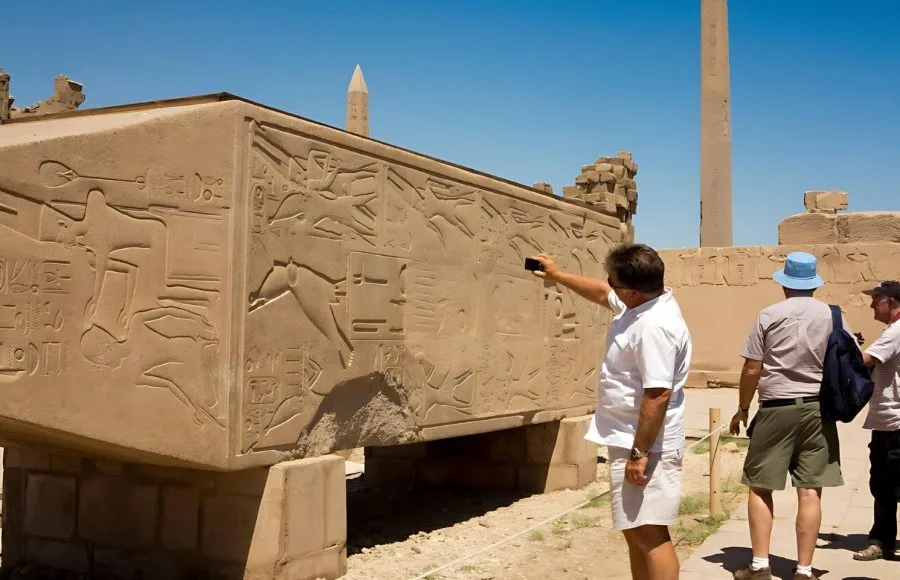 A tourist taking photos of Hapshetsut granite, specifically an engraved, massive block featuring detailed hieroglyphic reliefs and an obelisk in the background at Karnak Temple.