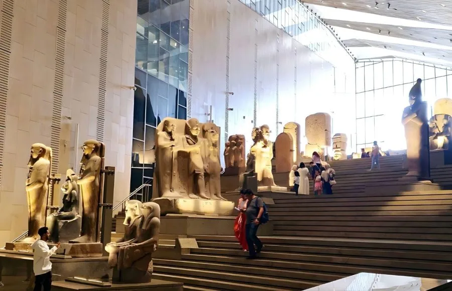 A high-angle interior shot inside GEM (The Grand Egyptian Museum, Giza), showing multiple tiers of sleek, modern display areas, stairs, and a central column of light with various ancient stone statues.