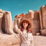 A smiling Girl Takes A selfie Against The Ruins of the Grandiose Karnak Temple, holding up her phone with colossal broken statues and columns in the sunlit background.