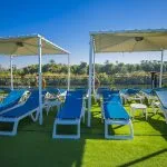 The sun deck of the Ms Blue Shadow iii Nile Cruise, showing rows of blue lounge chairs and white umbrellas on an artificial grass surface.