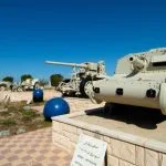 Military vehicles, including a Tank, artillery, and an armored car, are displayed outdoors at the El Alamein War Museum in Egypt under a bright blue sky.