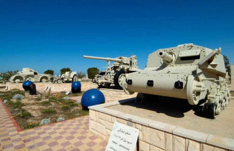 Military vehicles, including a Tank, artillery, and an armored car, are displayed outdoors at the El Alamein War Museum in Egypt under a bright blue sky.
