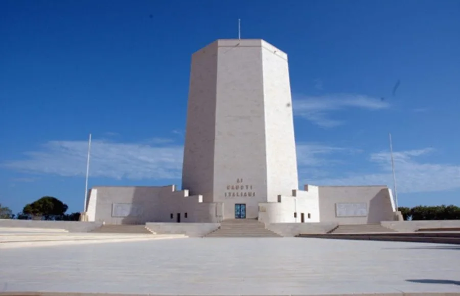 The large, octagonal white stone structure of the El Alamein Italian Memorial Building, standing under a clear blue sky and inscribed with "AI CADUTI ITALIANI" (To the Italian Fallen).