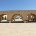 The arched stone entrance of the El Alamein Italian Memorial, with the words "SACRARIO MILITARE ITALIANO EL ALAMEIN" carved above the arches.
