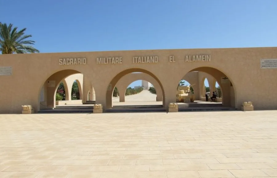 The arched stone entrance of the El Alamein Italian Memorial, with the words "SACRARIO MILITARE ITALIANO EL ALAMEIN" carved above the arches.