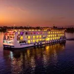 The illuminated HS Solaris I Nile Cruise ship glides along the Nile River at sunset, its lights reflecting beautifully on the water against a vibrant sky.