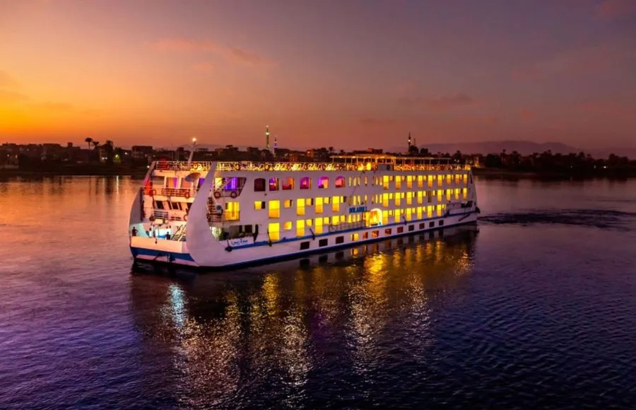 The illuminated HS Solaris I Nile Cruise ship glides along the Nile River at sunset, its lights reflecting beautifully on the water against a vibrant sky.