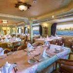 An elegantly set dining room on the HS Solaris I Nile Cruise, featuring beautifully prepared tables and large windows offering scenic views of the Nile River.