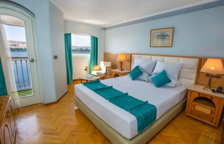 A bright and comfortable cabin on the HS Solaris I Nile Cruise, featuring a cozy bed, seating area, and a balcony door offering stunning views of the Nile River.