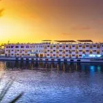 The HS Solaris I Nile Cruise ship beautifully illuminated at sunset, gracefully sailing on the calm waters of the Nile with a vibrant orange and purple sky.