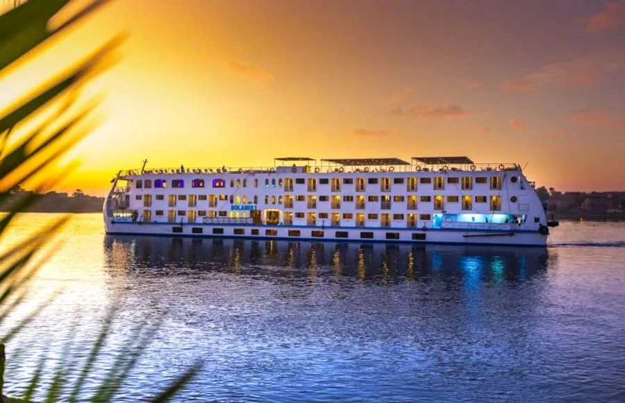 The HS Solaris I Nile Cruise ship beautifully illuminated at sunset, gracefully sailing on the calm waters of the Nile with a vibrant orange and purple sky.