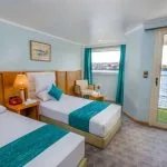 A well-appointed twin cabin on the HS Solaris I Nile Cruise, featuring two comfortable beds, a sitting area, and a balcony door with a beautiful view of the Nile River.