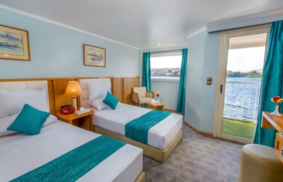 A well-appointed twin cabin on the HS Solaris I Nile Cruise, featuring two comfortable beds, a sitting area, and a balcony door with a beautiful view of the Nile River.