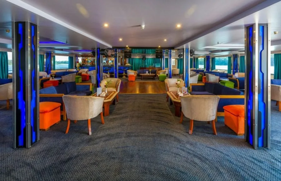 The vibrant lounge of the Solaris II Nile Cruise, featuring comfortable sofas, stylish lighting on columns, and wide windows with stunning Nile views