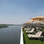 The sun deck of the Jaz Regent Nile Cruise, with lounge chairs and umbrellas facing a scenic view of the Nile River and lush green reeds.