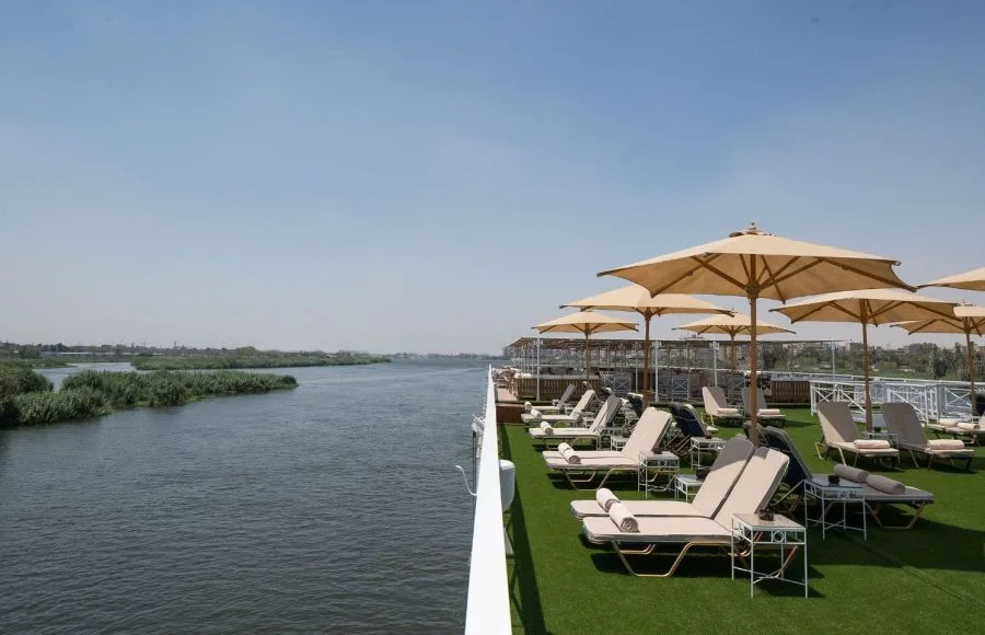 The sun deck of the Jaz Regent Nile Cruise, with lounge chairs and umbrellas facing a scenic view of the Nile River and lush green reeds.
