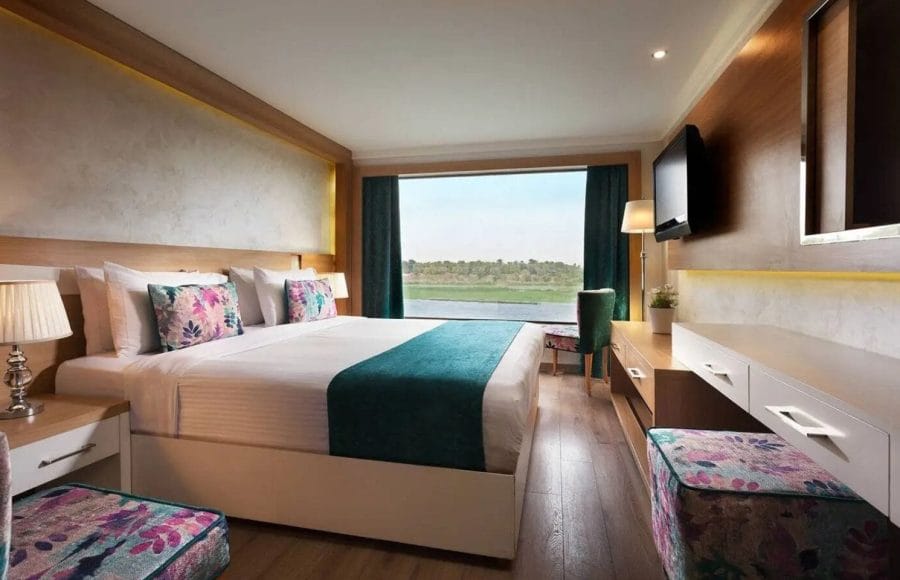 An elegant, wood-paneled hotel room on the MS Acamar Nile Cruise ship features twin beds, panoramic windows overlooking the Nile River, and modern furnishings.