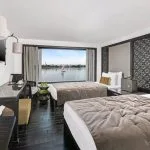 A modern twin-bedded cabin aboard the Movenpick MS Darakum Nile Cruise offers dark wood floors a stylish desk and a wide window view of a sailboat passing on the river