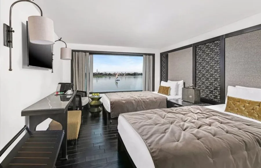 A modern twin-bedded cabin aboard the Movenpick MS Darakum Nile Cruise offers dark wood floors a stylish desk and a wide window view of a sailboat passing on the river