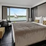 Luxurious suite aboard the Movenpick MS Darakum Nile Cruise offering a stunning Nile panorama with a traditional sailboat passing by for an unforgettable journey