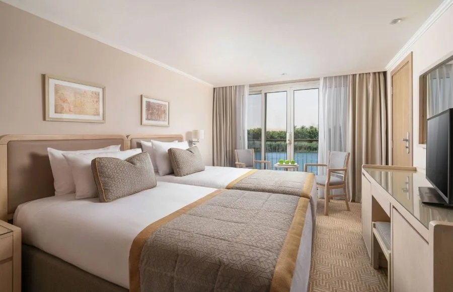 A clean and modern cabin with two twin beds on the MS Jaz Regent Nile Cruise, featuring a window and balcony overlooking the water.