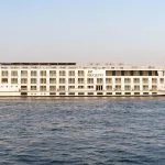 The MS Jaz Regent Nile Cruise ship sailing on the Nile River, with its multi-deck structure visible against the riverbanks.