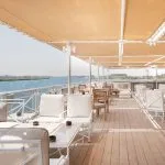 The sun deck lounge of the MS Jaz Regent Nile Cruise, with comfortable white sofas and chairs under a canopy, overlooking the serene Nile River.