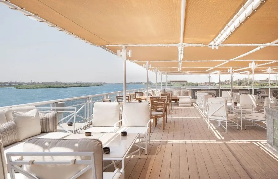 The sun deck lounge of the MS Jaz Regent Nile Cruise, with comfortable white sofas and chairs under a canopy, overlooking the serene Nile River.