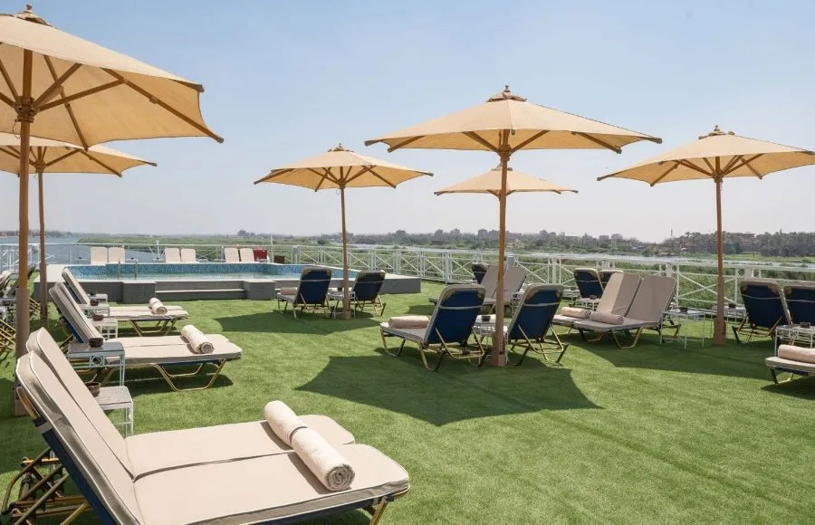 The expansive sun deck of the MS Jaz Regent Nile Cruise, featuring a small pool, numerous lounge chairs with umbrellas, and a view of the Nile River banks.
