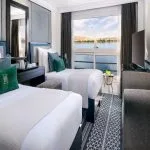 A twin-bed cabin on the Movenpick Hamees Nile Cruise features modern decor, comfortable bedding, and a large panoramic window overlooking the Nile River.