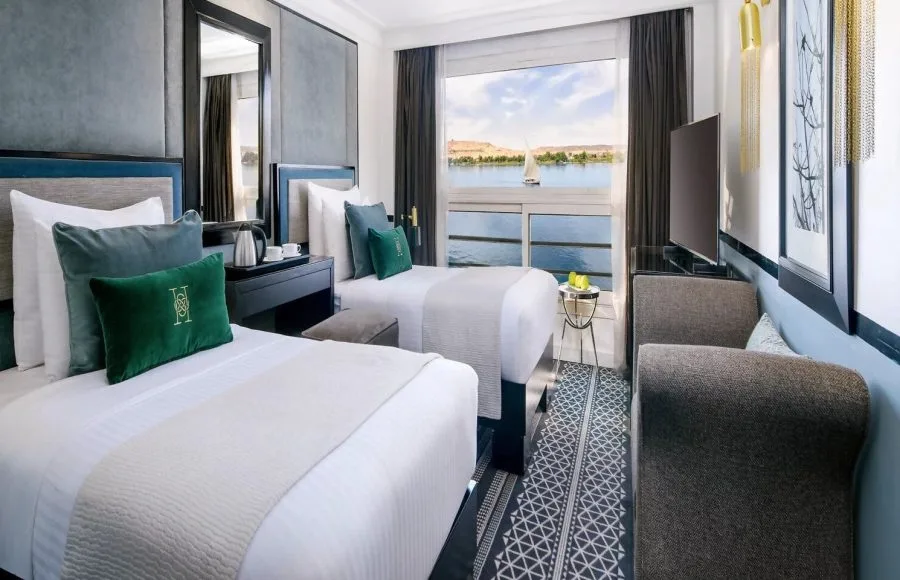 A twin-bed cabin on the Movenpick Hamees Nile Cruise features modern decor, comfortable bedding, and a large panoramic window overlooking the Nile River.