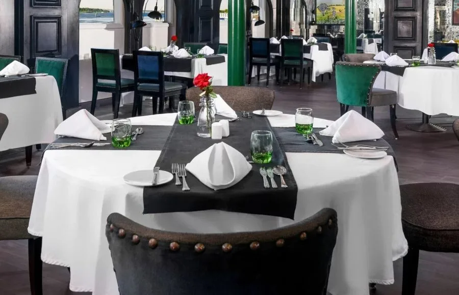 Elegant dining setup inside the Movenpick Hamees Nile Cruise restaurant featuring crisp white linens, deep green accents, and a red rose centerpiece awaiting guests.
