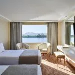 A brightly lit Mövenpick MS Royal Lotus Nile Cruise cabin with twin beds and a panoramic window overlooking the scenic Nile River.