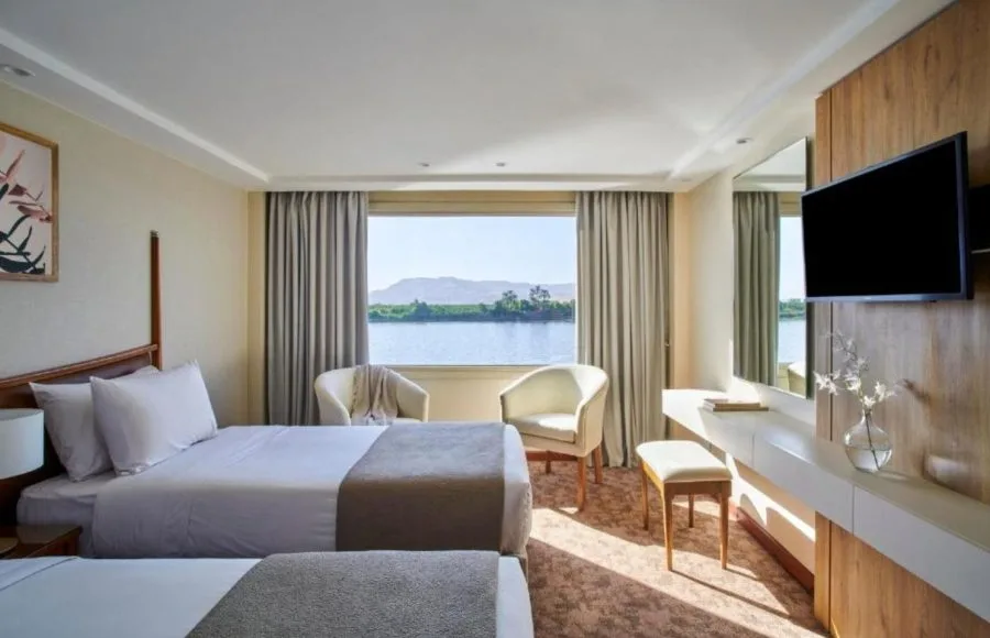 A brightly lit Mövenpick MS Royal Lotus Nile Cruise cabin with twin beds and a panoramic window overlooking the scenic Nile River.