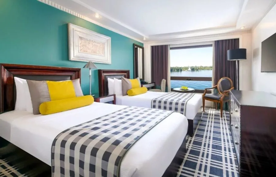 A twin-bed cabin on the Movenpick MS Sunray featuring colorful decor and a large window overlooking the Nile River.