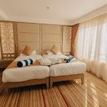 A brightly room with two twin beds, featuring beige upholstered headboards with decorative wooden screens on either side, and large windows covered by sheer white and heavy tan curtains.