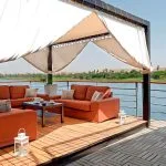 A shaded outdoor seating area with comfortable sofas and stunning river views on the sundeck of the ms nile premium cruise.